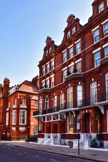 A street view of a multi-storey red brick building with large sash windows, some featuring small balconies with black wrought iron railings, and white decorative elements under the roofline. The pavem