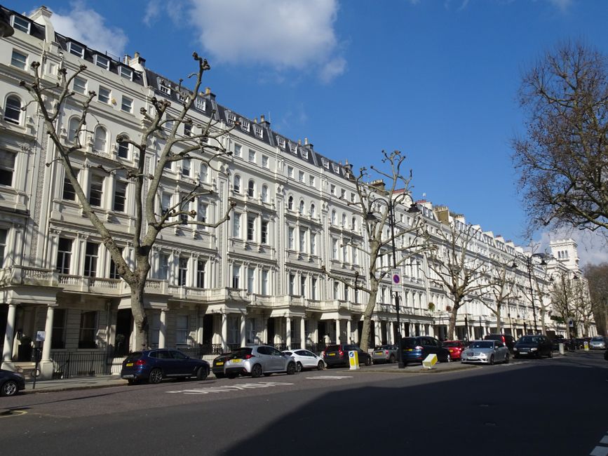 A street view of a multi-storey red brick building with large sash windows, some featuring small balconies with black wrought iron railings, and white decorative elements under the roofline. The pavem