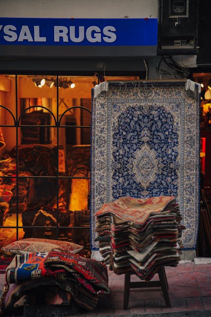 A row of colorful, intricately patterned rugs hanging on a stone brick wall along a paved street. The rugs feature traditional geometric and floral designs in shades of red, orange, beige, and blue. T