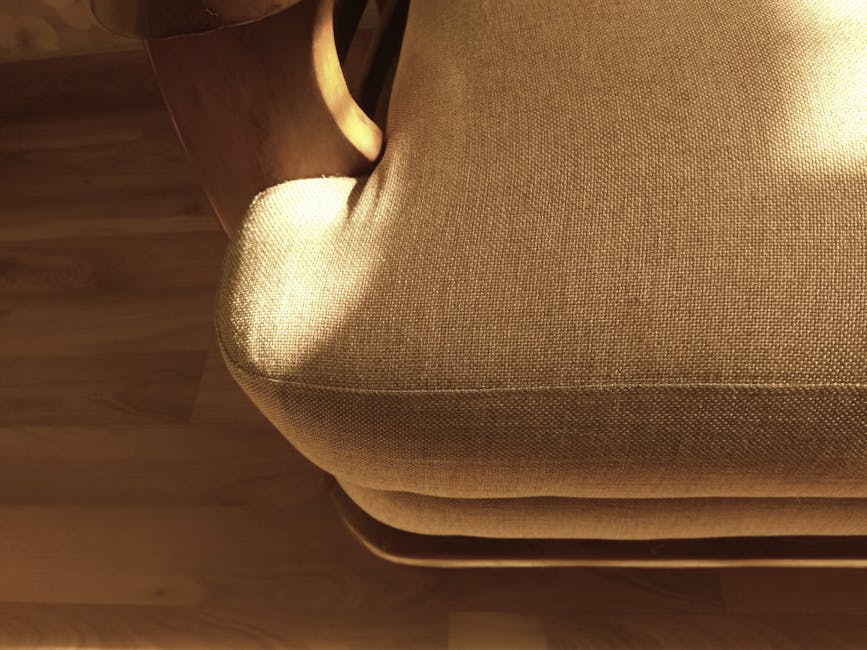Close-up view of a beige upholstered armchair with textured fabric, situated on a wooden floor in a well-lit room. The cushion appears clean and well-maintained, with subtle shadows cast by a nearby l